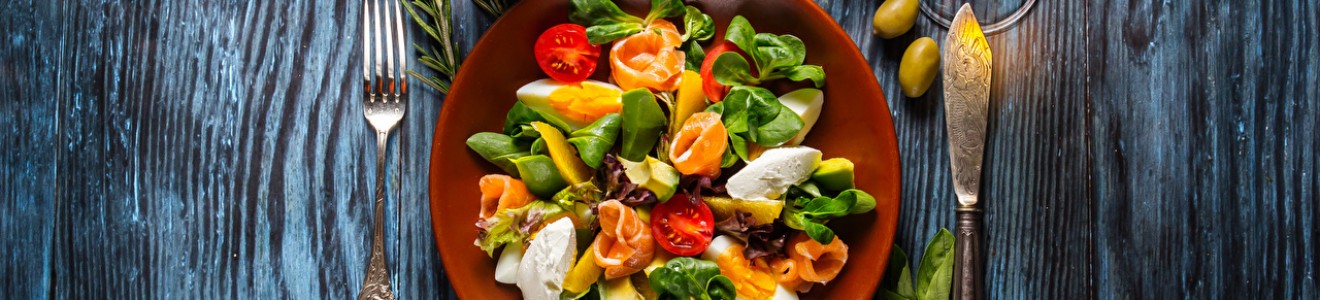 A fresh salad with greens, boiled eggs, cherry tomatoes, and smoked salmon arranged on a brown plate surrounded by herbs, spices, and a glass of amber-colored drink on a wooden table.