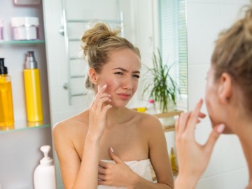 A woman wrapped in a towel examines and touches acne on her cheek while looking in a bathroom mirror.