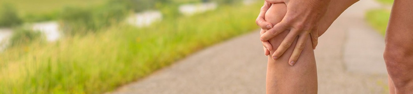 Person in athletic clothing holding their knee on a paved path with greenery and hills in the background.