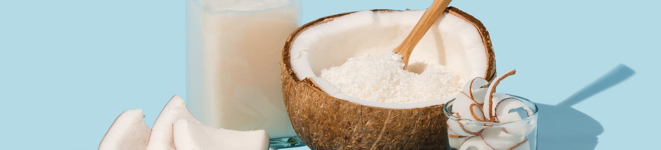 A bottle of coconut milk, fresh coconut slices on a plate, a halved coconut filled with shredded coconut and a wooden spoon, and a glass with coconut curls on a blue background.