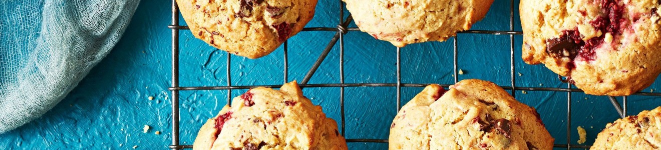 Freshly baked chocolate chip and raspberry cookies cooling on a wire rack over a blue textured surface.