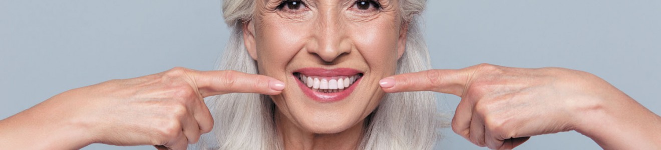 A smiling older woman with gray hair points to her teeth against a light gray background.