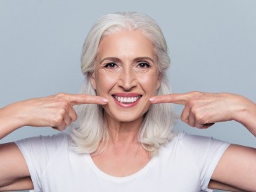 A smiling older woman with gray hair points to her teeth against a light gray background.