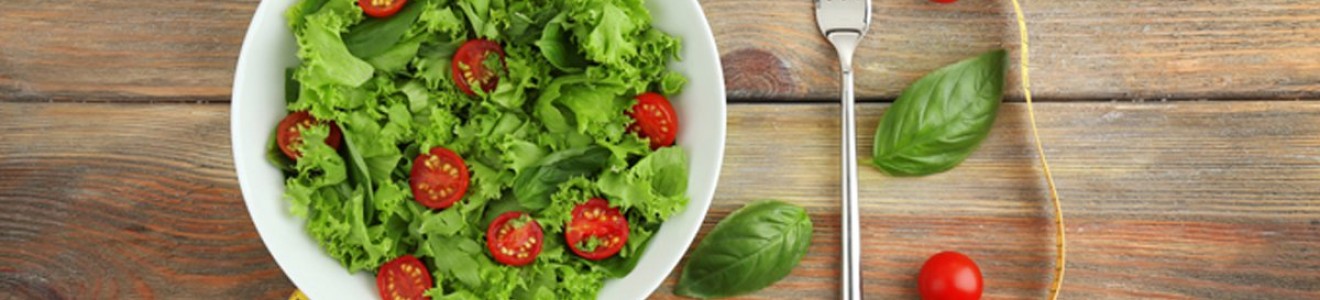A bowl of fresh green lettuce and cherry tomato salad with a fork, basil leaves, and a yellow measuring tape on a wooden surface.