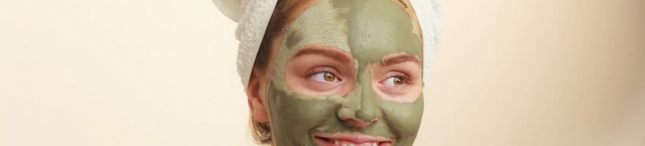 A woman with a green facial mask and a white towel wrapped around her head smiling and looking to the side.