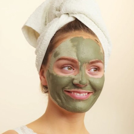 A woman with a green facial mask and a white towel wrapped around her head smiling and looking to the side.