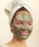 A woman with a green facial mask and a white towel wrapped around her head smiling and looking to the side.