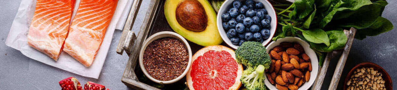 A selection of fresh healthy foods including salmon, avocado, blueberries, almonds, spinach, broccoli, apples, grapefruit, flaxseeds, pomegranate, and various seeds and spices.