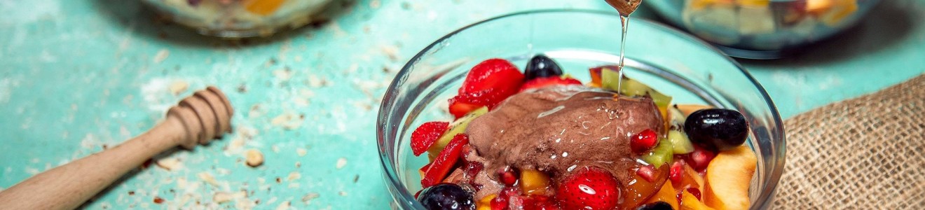 Three glass bowls filled with mixed fruit and yogurt, with honey being drizzled onto one bowl.