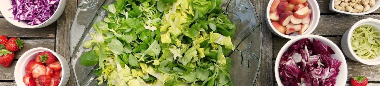 A glass bowl of mixed greens surrounded by small bowls of various fresh fruits, vegetables, nuts, and mushrooms on a wooden table.