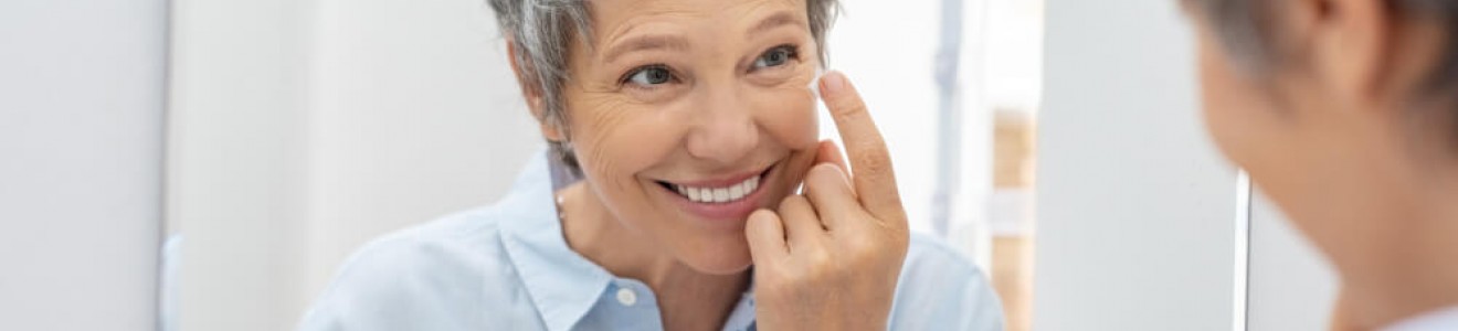 A smiling middle-aged woman with short gray hair applies cream to her face while looking in a mirror.