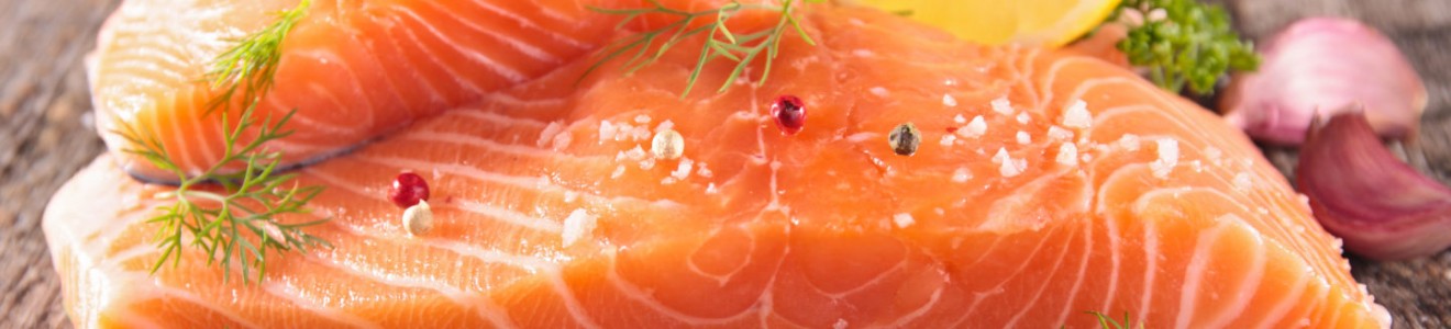 Raw salmon fillets seasoned with coarse salt, peppercorns, fresh dill, parsley, and a lemon wedge on a wooden surface.