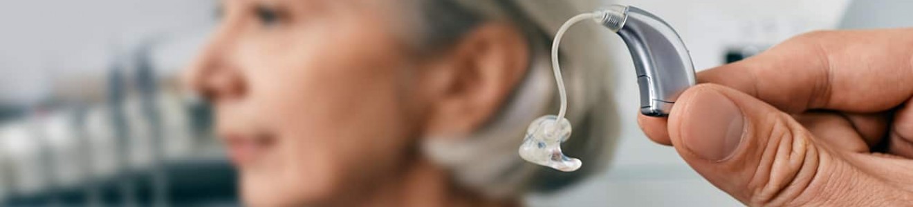 Hand holding a hearing aid near an elderly woman with gray hair in a clinical setting.