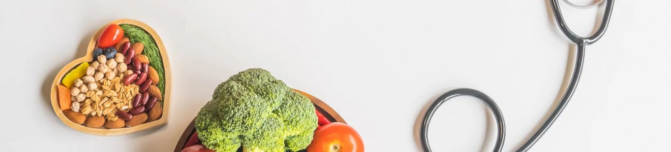 A stethoscope with a red heart, a bowl of fresh vegetables and fruits, and a heart-shaped dish of nuts and seeds on a white background.