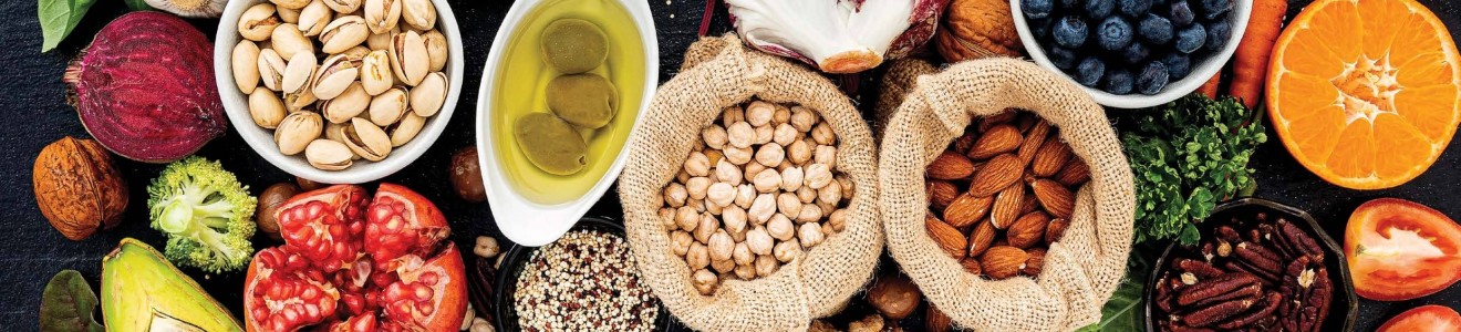 A variety of fresh fruits, vegetables, nuts, seeds, and olive oil arranged on a dark surface.