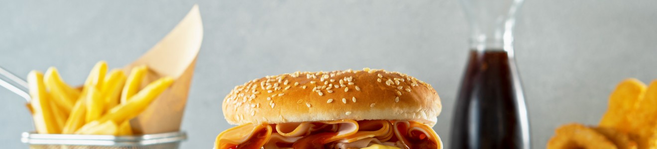 A fried chicken sandwich with lettuce, tomato, cheese, and sauce on a sesame bun, accompanied by French fries, onion rings, and a glass bottle of soda.
