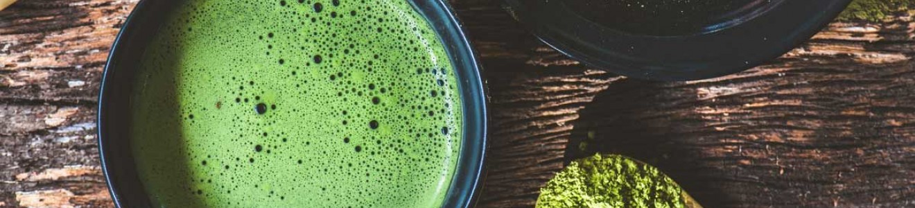 A cup of frothy matcha tea, a wooden spoon with matcha powder, a plate with matcha powder, and a bamboo whisk on a rustic wooden surface.