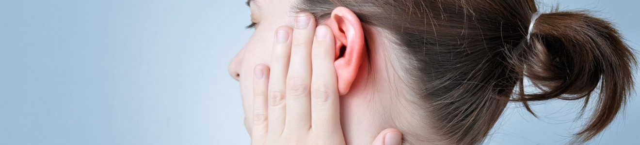 A woman with a ponytail is holding her red, irritated ear with her hand against a light blue background.