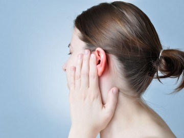 A woman with a ponytail is holding her red, irritated ear with her hand against a light blue background.