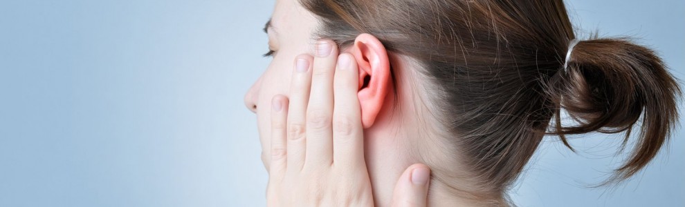 A woman with a ponytail is holding her red, irritated ear with her hand against a light blue background.