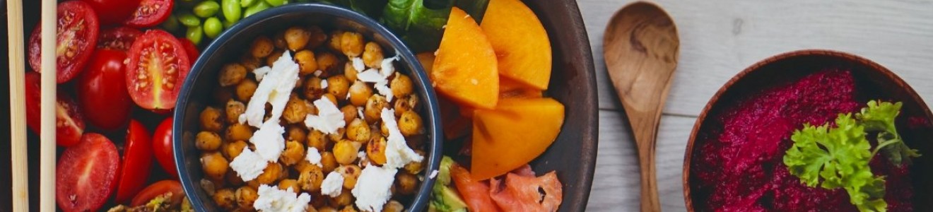 A colorful Buddha bowl with chickpeas, avocado, cherry tomatoes, spinach, persimmons, smoked salmon, and edamame, with chopsticks and a bowl of beet hummus on the side.