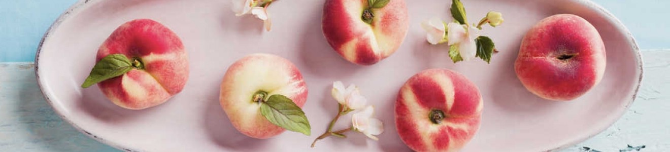 A pink oval plate holding six flat peaches with green leaves and small white flowers, set on a light blue surface.