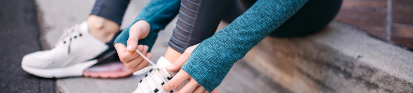 Person sitting on a curb tying the laces of white athletic shoes while wearing black leggings and a teal long-sleeve top.