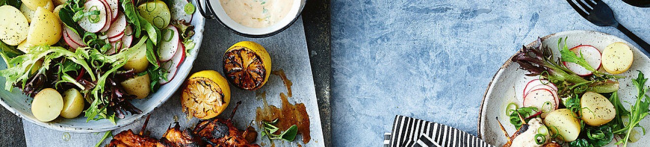 Grilled chicken skewers served with a potato and radish salad, grilled lemons, and a creamy dipping sauce on a blue textured background.