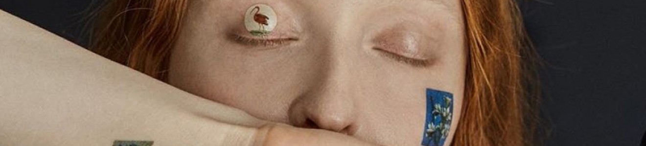 A person with red hair has their eyes closed and arms raised, with various small colorful stickers or tattoos of animals and plants on their face, arms, and neck.