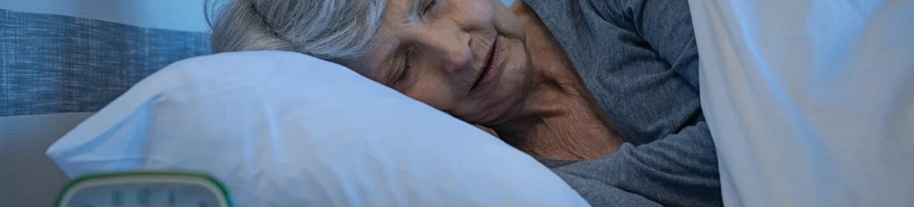 An elderly woman with gray hair is sleeping on her side in bed, covered with a white blanket and resting on a white pillow, with an alarm clock on a bedside table.