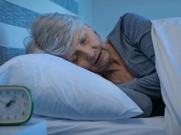 An elderly woman with gray hair is sleeping on her side in bed, covered with a white blanket and resting on a white pillow, with an alarm clock on a bedside table.