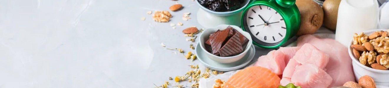 A variety of healthy foods including salmon, nuts, kiwi, oatmeal, milk, chocolate, and prunes arranged next to a green alarm clock on a white surface.
