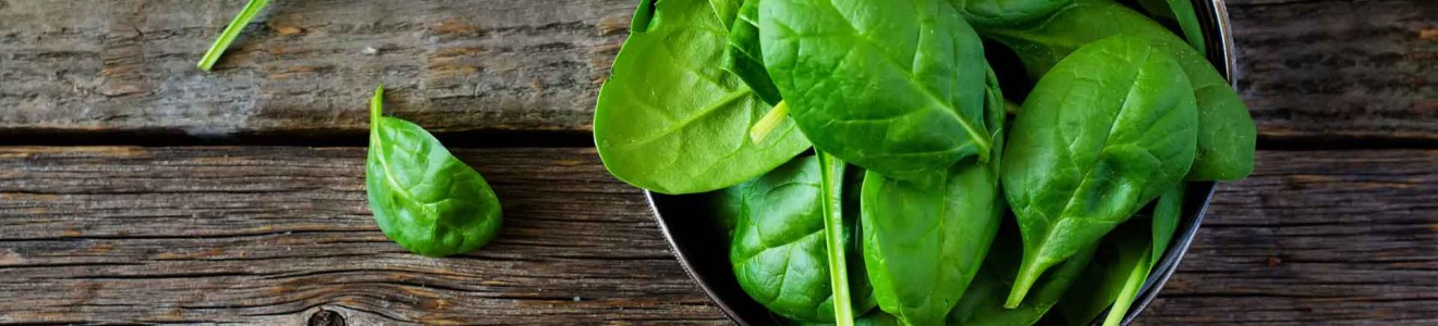 Fresh spinach leaves in a black bowl on a rustic wooden surface with two leaves beside the bowl.