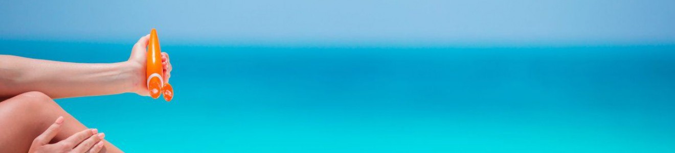 Person applying sunscreen on their leg at a sunny beach with clear blue ocean in the background.