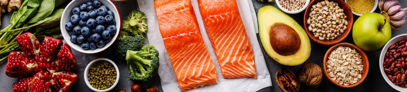 Two raw salmon fillets surrounded by various healthy foods including fruits, vegetables, nuts, seeds, and spices on a gray surface.
