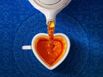 A top-down view of tea being poured from a white teapot into a heart-shaped cup on a blue patterned surface.