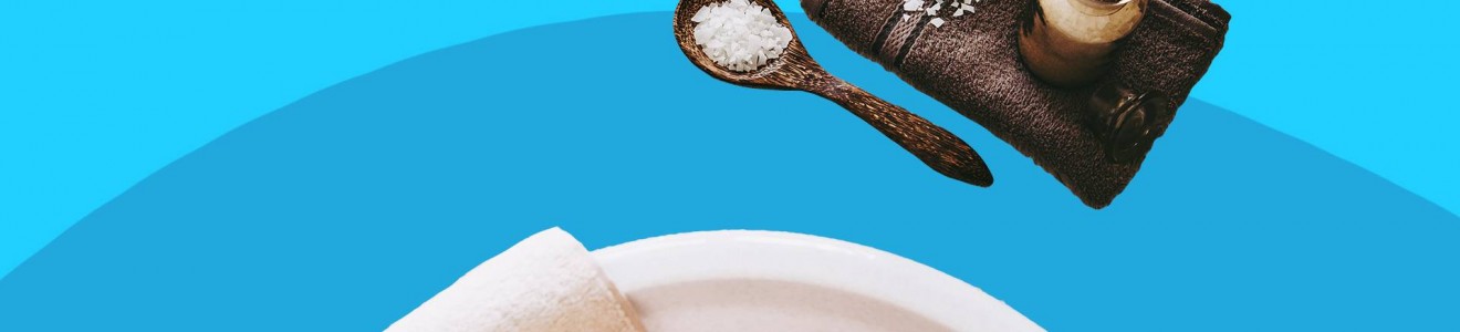 Feet soaking in a foot bath with a towel, exfoliating stone, bath salts, and a jar of treatment on a blue background.