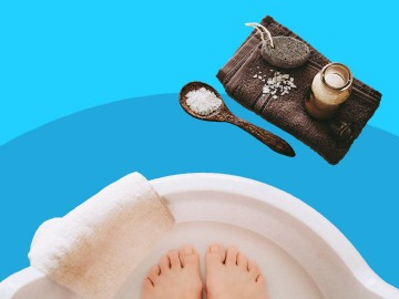 Feet soaking in a foot bath with a towel, exfoliating stone, bath salts, and a jar of treatment on a blue background.