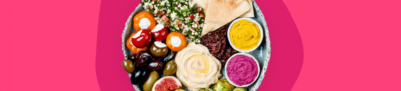 A vibrant Mediterranean platter featuring hummus, pita bread, falafel, stuffed mini peppers, olives, tabbouleh, fig halves, two dips, and a pepper, set against a bright pink background.