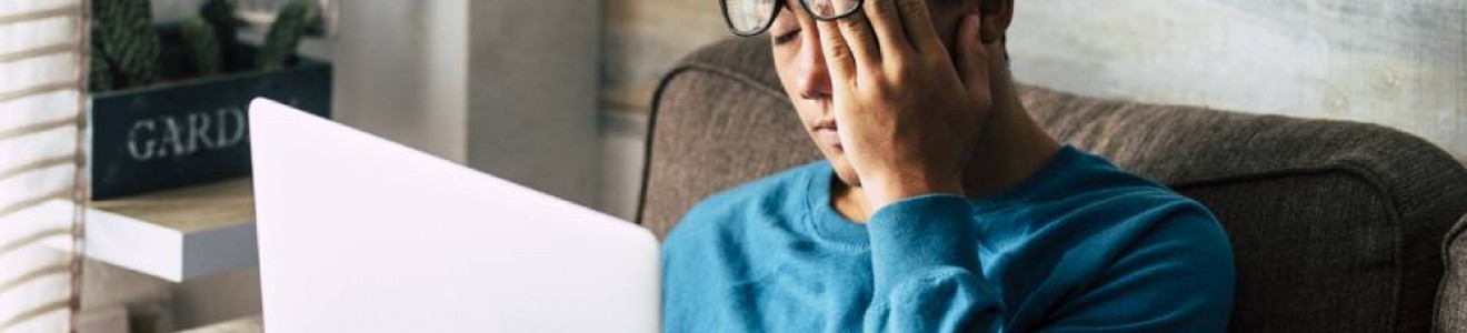 A young man sitting on a couch with a laptop on his lap, rubbing his eyes through his glasses, appearing tired or stressed.