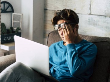 A young man sitting on a couch with a laptop on his lap, rubbing his eyes through his glasses, appearing tired or stressed.