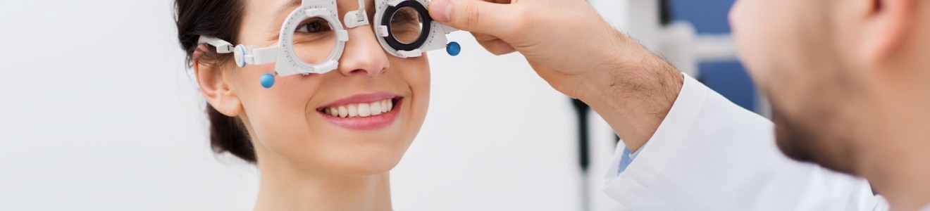 A woman smiling during an eye exam as an optometrist adjusts a phoropter on her face.